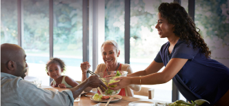 Imagem de uma família reunida na mesa de jantar, onde uma mulher está levantada servindo uma tijela de salada a um homem