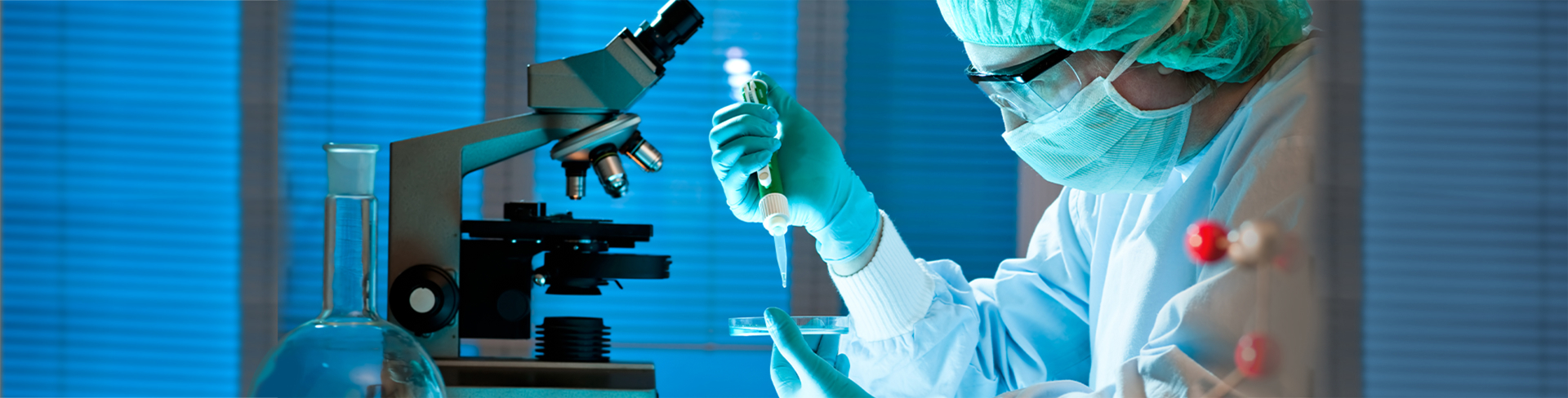 cientista vestindo um avental, máscara, toca e luvas, fazendo análise de composição em um acrílico com uma seringa, em um laboratório contendo um microscópio